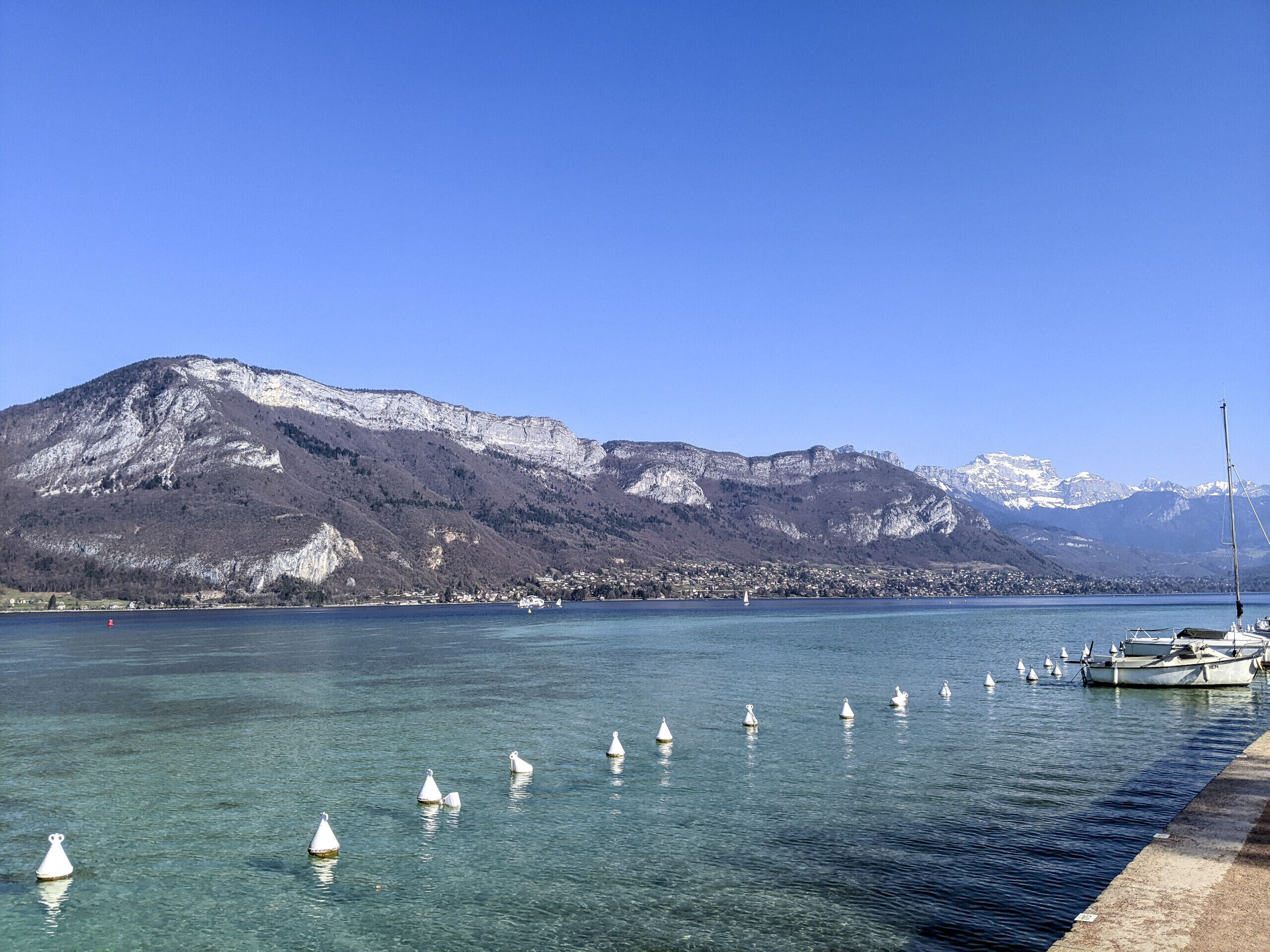 A clear lake with buoys, a boat docked on the right, and a mountain range in the background.