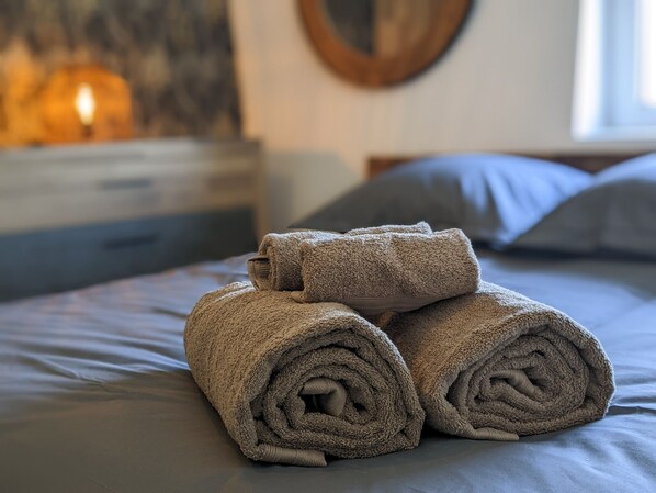 A neatly made bed with rolled towels, a wooden headboard, and a wall-mounted circular mirror.