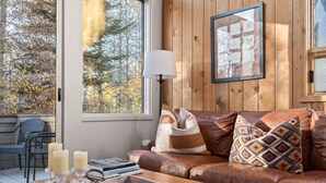 Ein gemütliches Wohnzimmer mit einer Holzvertäfelung, großen Fenstern, einem Ledersofa und einem Couchtisch mit Büchern und Kerzen.