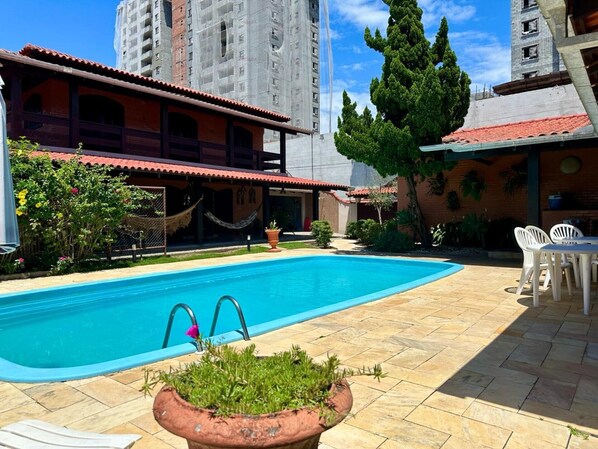 Una piscina con solárium de baldosas, rodeada por un edificio con techo de tejas rojas y un árbol grande.