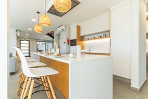 Une cuisine moderne avec des meubles blancs, un îlot central doté de tabourets de bar, un éclairage suspendu et une vue sur l’extérieur.
