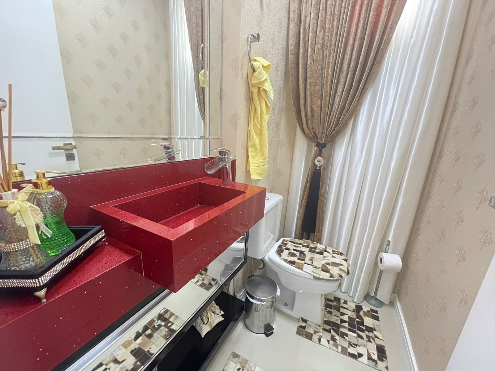 Un baño pequeño con un lavabo rojo, un inodoro con asiento estampado, un espejo y un cubo de basura.
