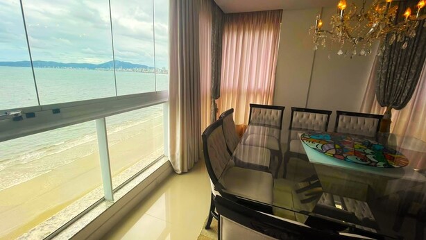 A dining area with a glass table and chairs, a chandelier, and a view of the beach and city skyline through large windows. - Cobertura Infinity Blue - Itapema-SC (Itapema)