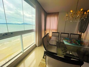 Un comedor con una mesa y sillas de vidrio, una araña y vistas a la playa y el horizonte urbano a través de amplios ventanales.