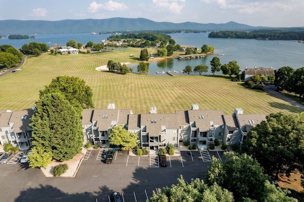 A residential area with a large grassy field, a parking lot, and a body of water in the background. - Tranquility Cove at Smith Mountain Lake —Your Home Away From Home! (Moneta)