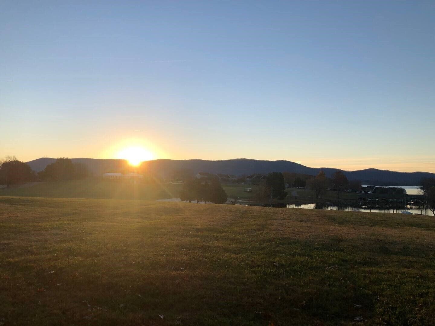 Auringonlasku niityn yllä, taustalla näkyy vesistö ja kukkulat.