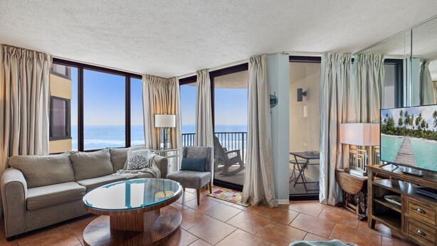 A spacious living room with a large window overlooking the ocean, a round coffee table, a sofa, a chair, and a television. - Lands' End @ Sunbird :5th floor Sleeps 4 Gulf Front (Panama City)