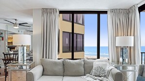 A modern living room with a large window offering a view of the beach, a sofa, a coffee table, and a dining area with chairs and a lamp. - Lands' End @ Sunbird :5th floor Sleeps 4 Gulf Front (Panama City)