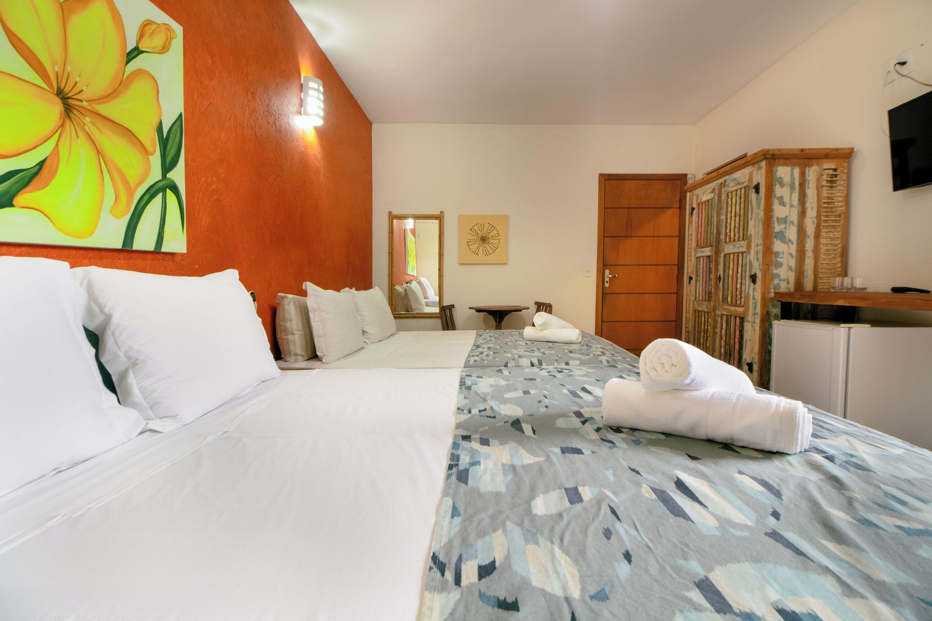 A hotel room with a bed, a large floral painting, a wooden wardrobe, and a small table with a mirror.