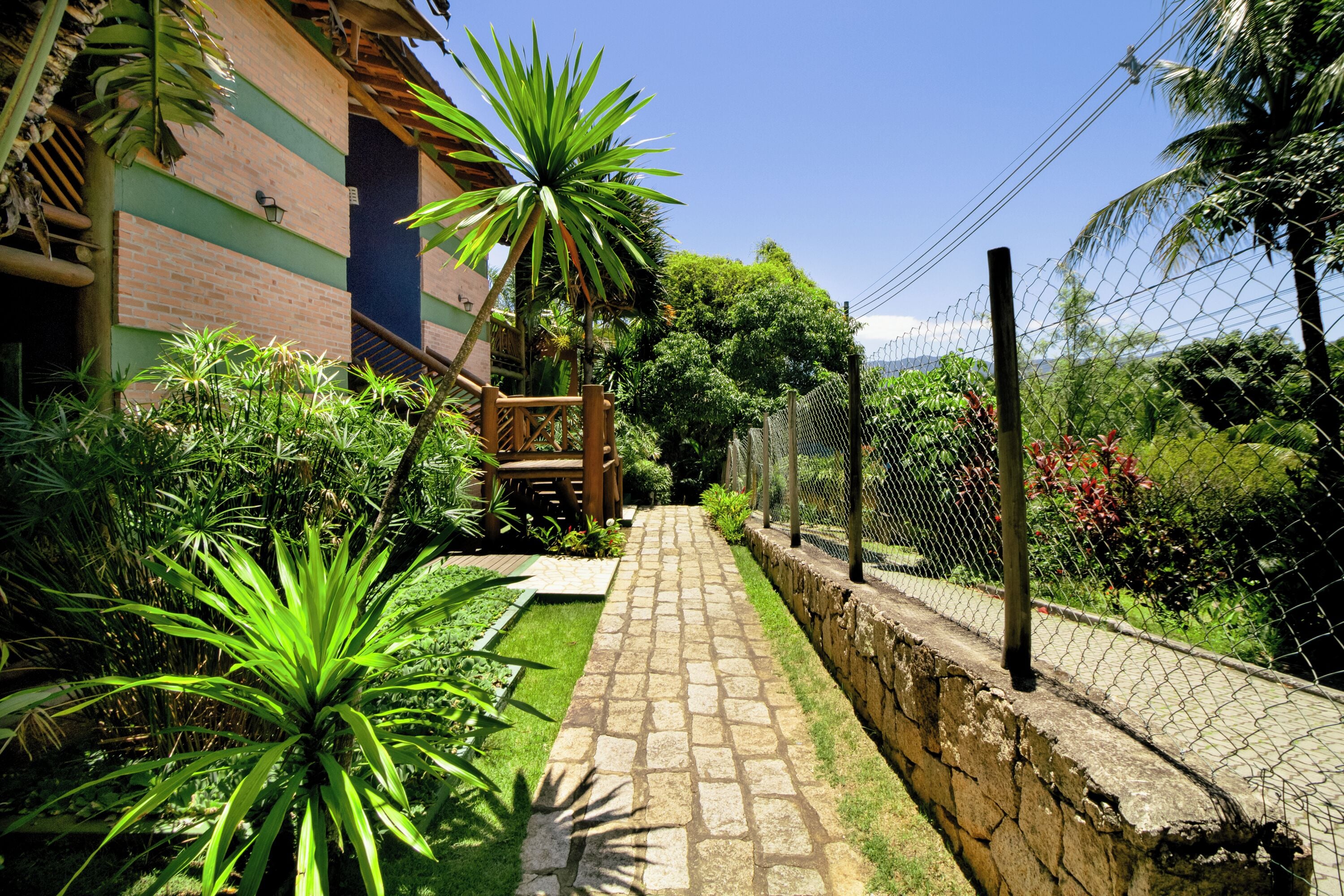 A paved pathway leading to a wooden deck, surrounded by lush greenery and a chain-link fence.