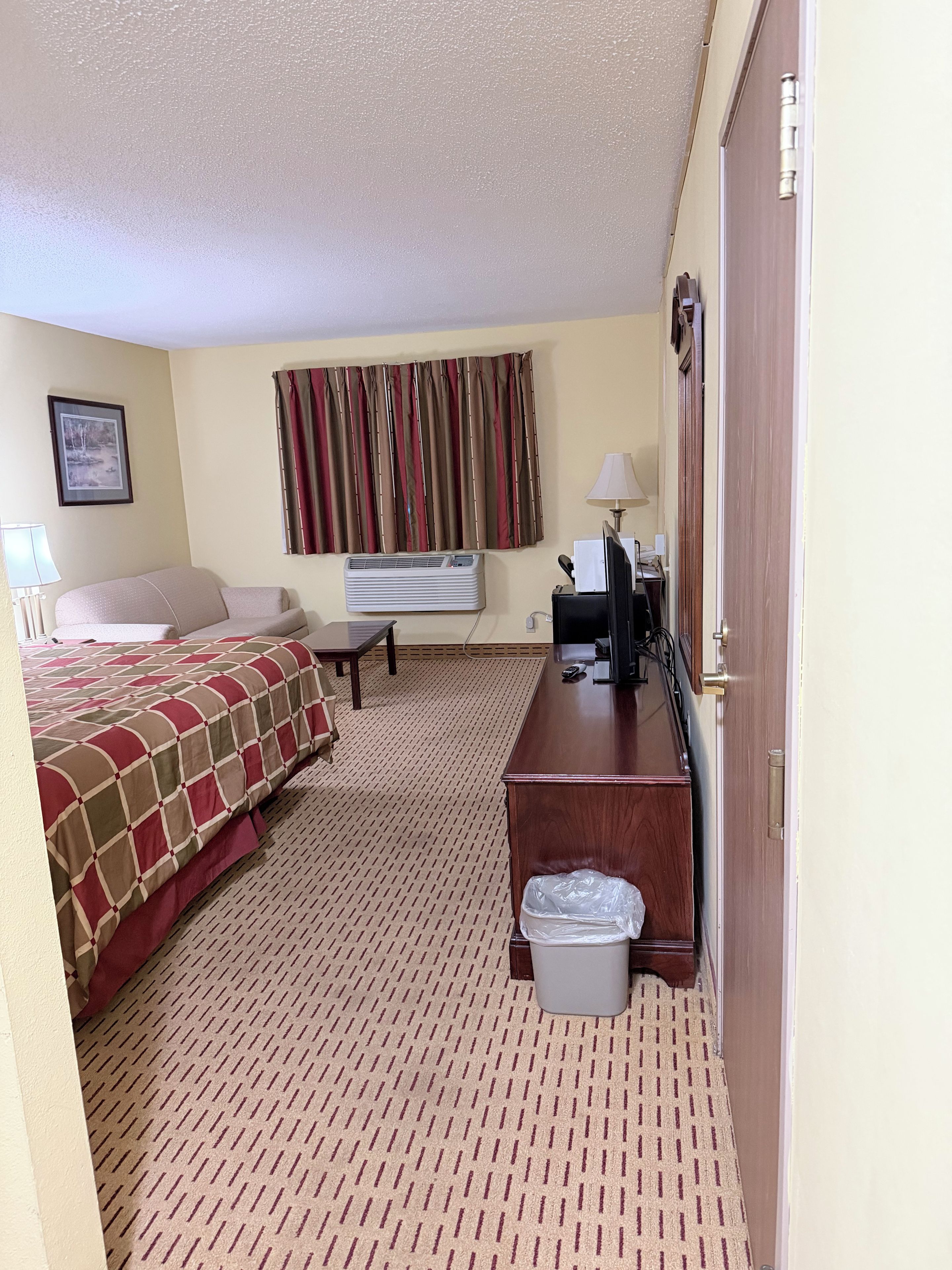 A hotel room with a bed, a desk, a chair, a waste bin, a lamp, and a window with curtains.