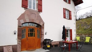 Ein Gebäude mit Holztür, Steinbogen und roten Fensterläden. Ein Grill und Sitzgelegenheiten mit Stühlen sind vorhanden.