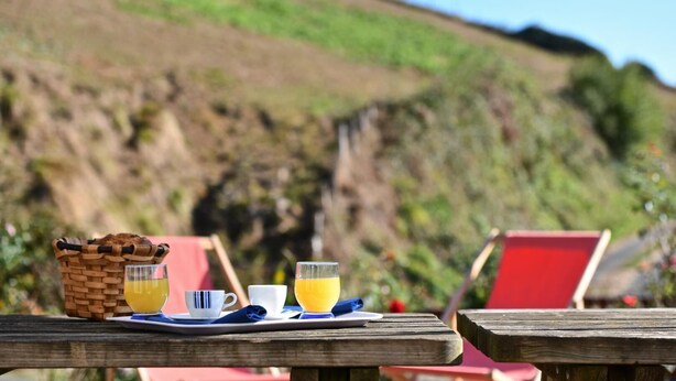 A breakfast setup with a basket, two cups, and a glass of orange juice on a wooden table with a red chair in the background. - SARAHANDIA - ALDUDES (LES-ALDUDES)