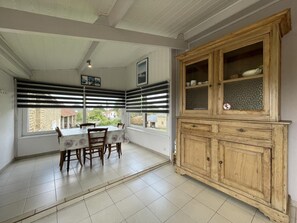 Een eethoek met een tafel, stoelen, een houten kast en grote ramen met jaloezieën.