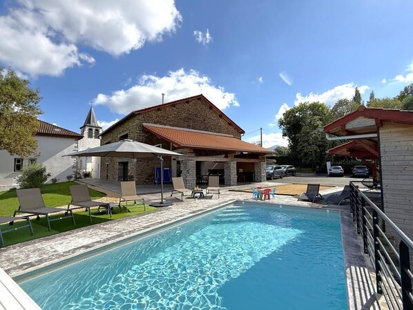 Une piscine avec des chaises longues et des parasols, un bâtiment en pierre avec un toit rouge et une église dotée d’un clocher.