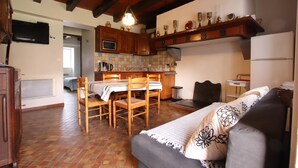 A cozy living room with a sofa, a dining table with chairs, a kitchen area with wooden cabinets and a microwave, and a TV mounted on the wall. - Caricaburia - Juxue - Gîte (JUXUE)