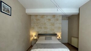 Une chambre avec un mur en pierre, un lit agrémenté d’un coussin orné du mot « NATURE », deux lampes de chevet et un banc en bois.