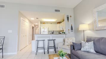 A modern living room with a grey sofa, a coffee table with remote controls, and a kitchen area in the background.