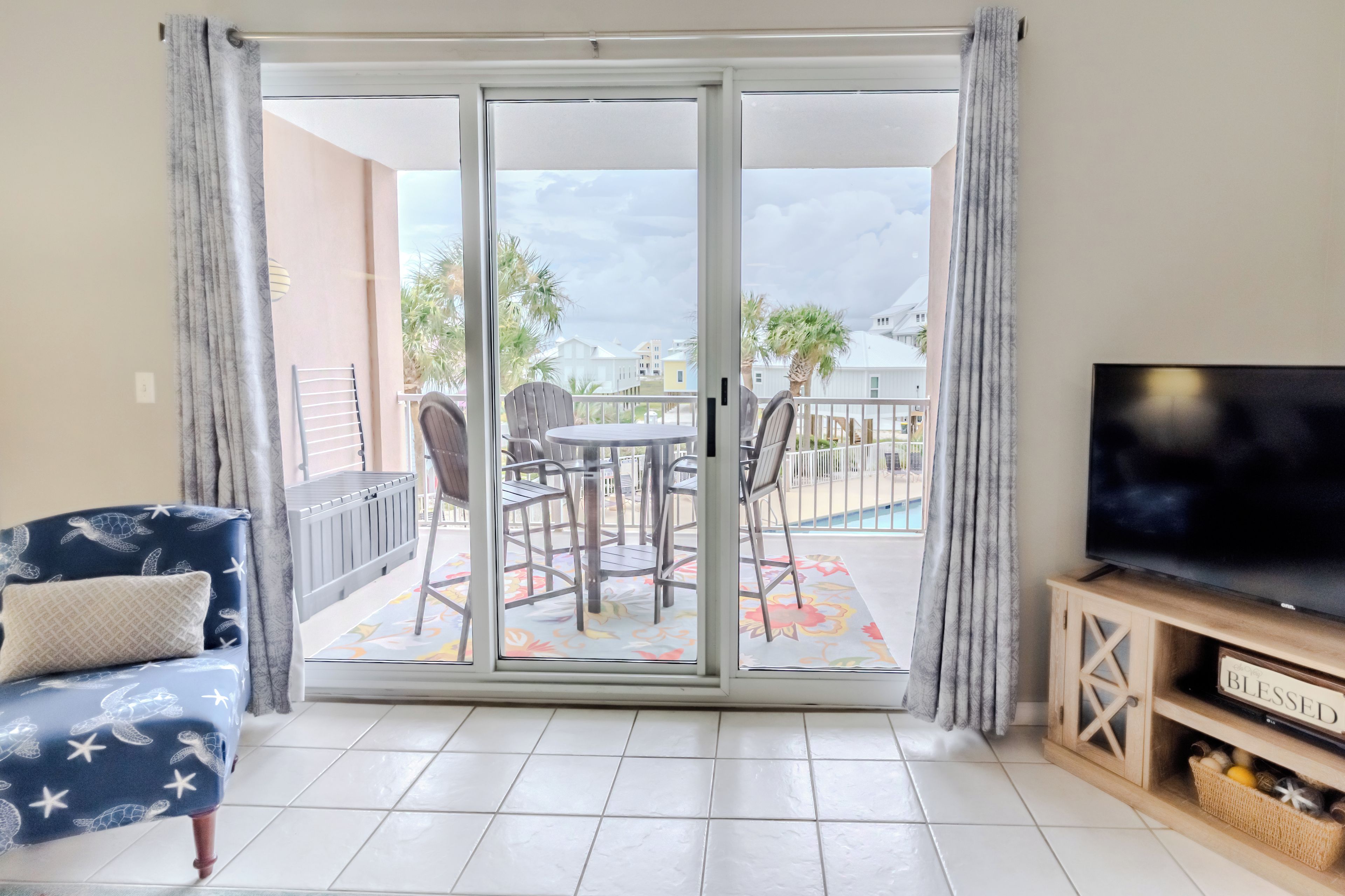 A living room with a sofa, a television, and a sliding glass door leading to a balcony with a pool view.