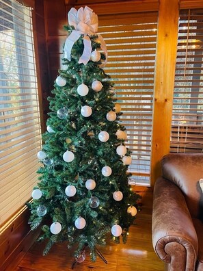 A Christmas tree with white ornaments and a large bow, placed in a corner with wooden blinds and a leather sofa.