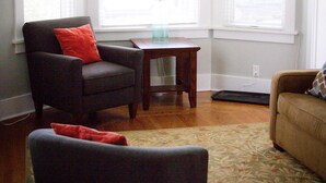 A living room with a dark armchair, a small wooden table, and a beige sofa. There are large windows with white blinds and a ceiling fan. - Historic Home Near Downtown & UW (Laramie)