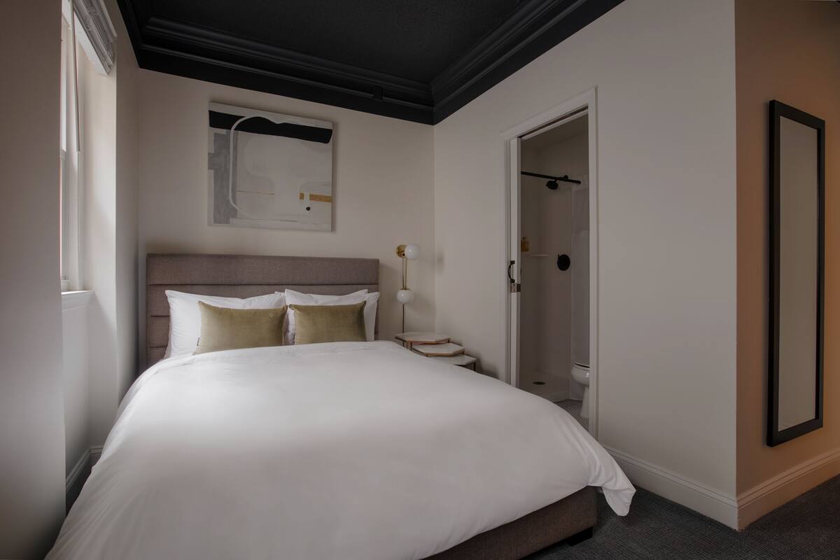 A hotel room with a large bed, a bedside table, a nightstand, a wall-mounted artwork, a door to a bathroom, and a mirror on the wall.