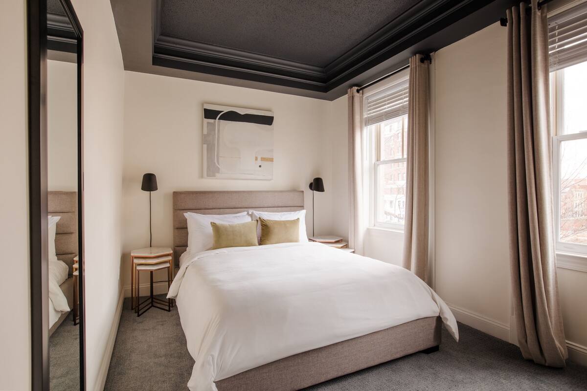 A hotel room with a large bed, a bedside table, a nightstand, a wall-mounted artwork, and a window with curtains.