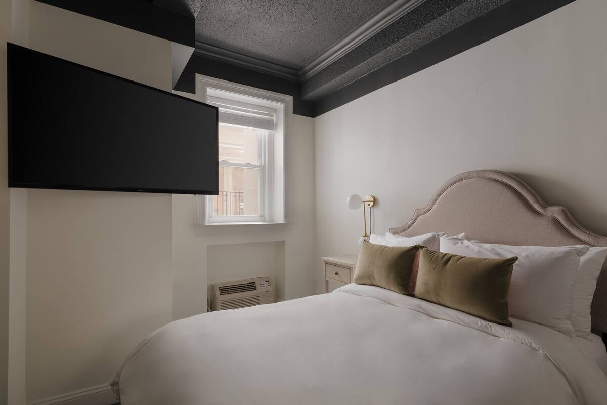 A bedroom with a bed, a nightstand, a lamp, a window, and a mounted TV.