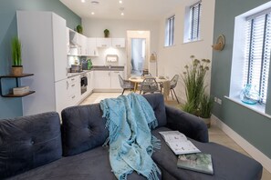 A modern living room with a sofa, a dining table with chairs, a kitchen area, and a window with blinds. - Courtyard Retreat (Southampton)