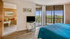 A bedroom with a bed, a television on a dresser, a balcony with chairs, and a view of the landscape. - Hibiscus Resort Condo with Ocean View (St. Augustine)