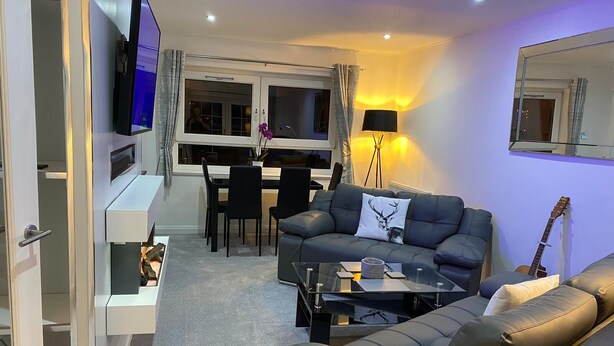 Ett modernt vardagsrum med en grå soffa, ett soffbord och en tv som är monterad på väggen. Det finns också en matplats med ett bord och stolar, och en golvlampa.