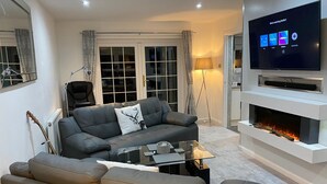 Ett modernt vardagsrum med en grå soffa, ett soffbord i glas, en platt-TV som är monterad på en vit vägg och en öppen spis.