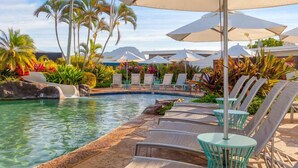 Un área junto a la piscina con sillones de descanso, sombrillas y plantas tropicales.