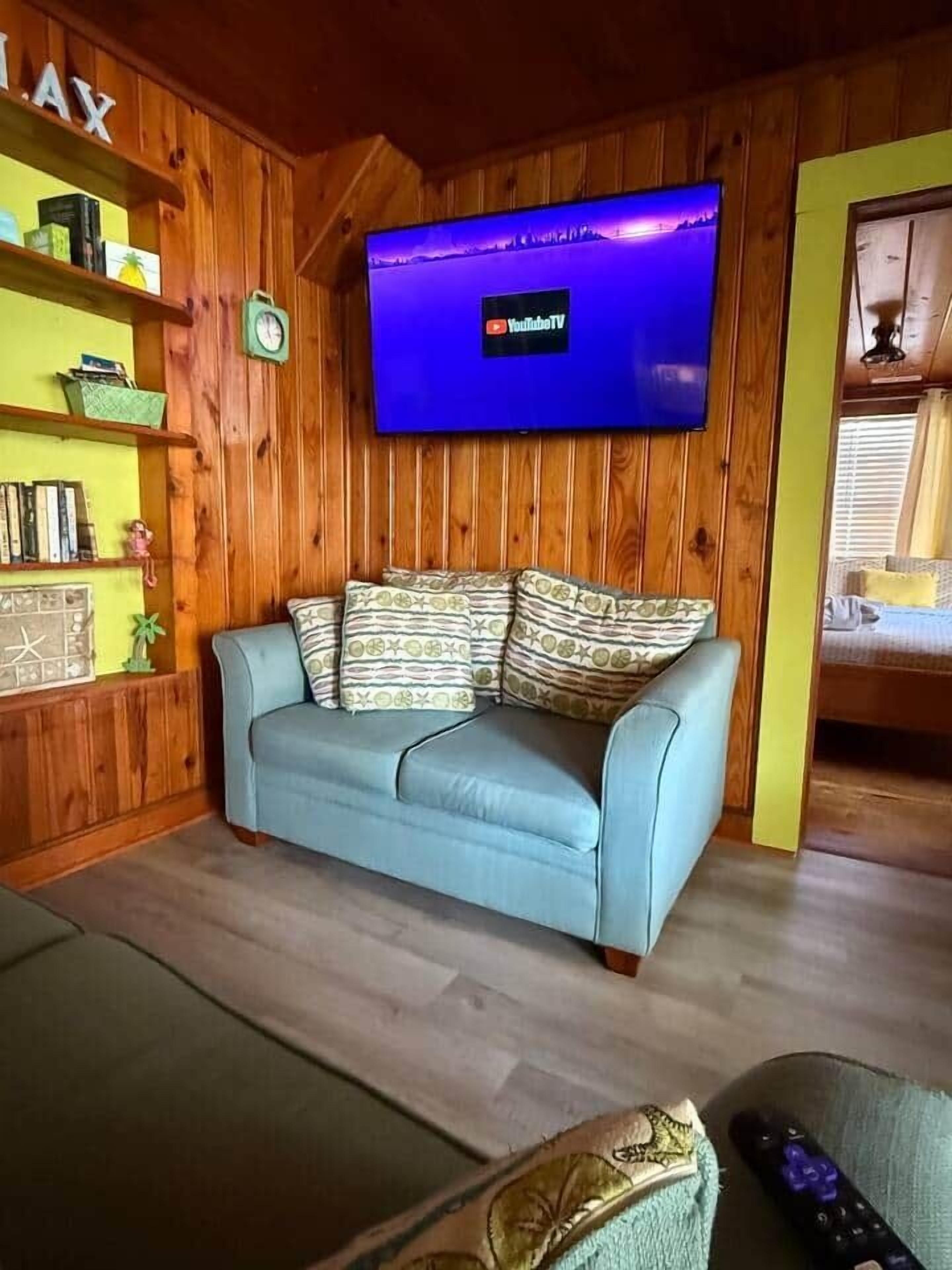 A living room with a blue sofa, a flat-screen TV mounted on the wall, wooden paneling, and a bookshelf.