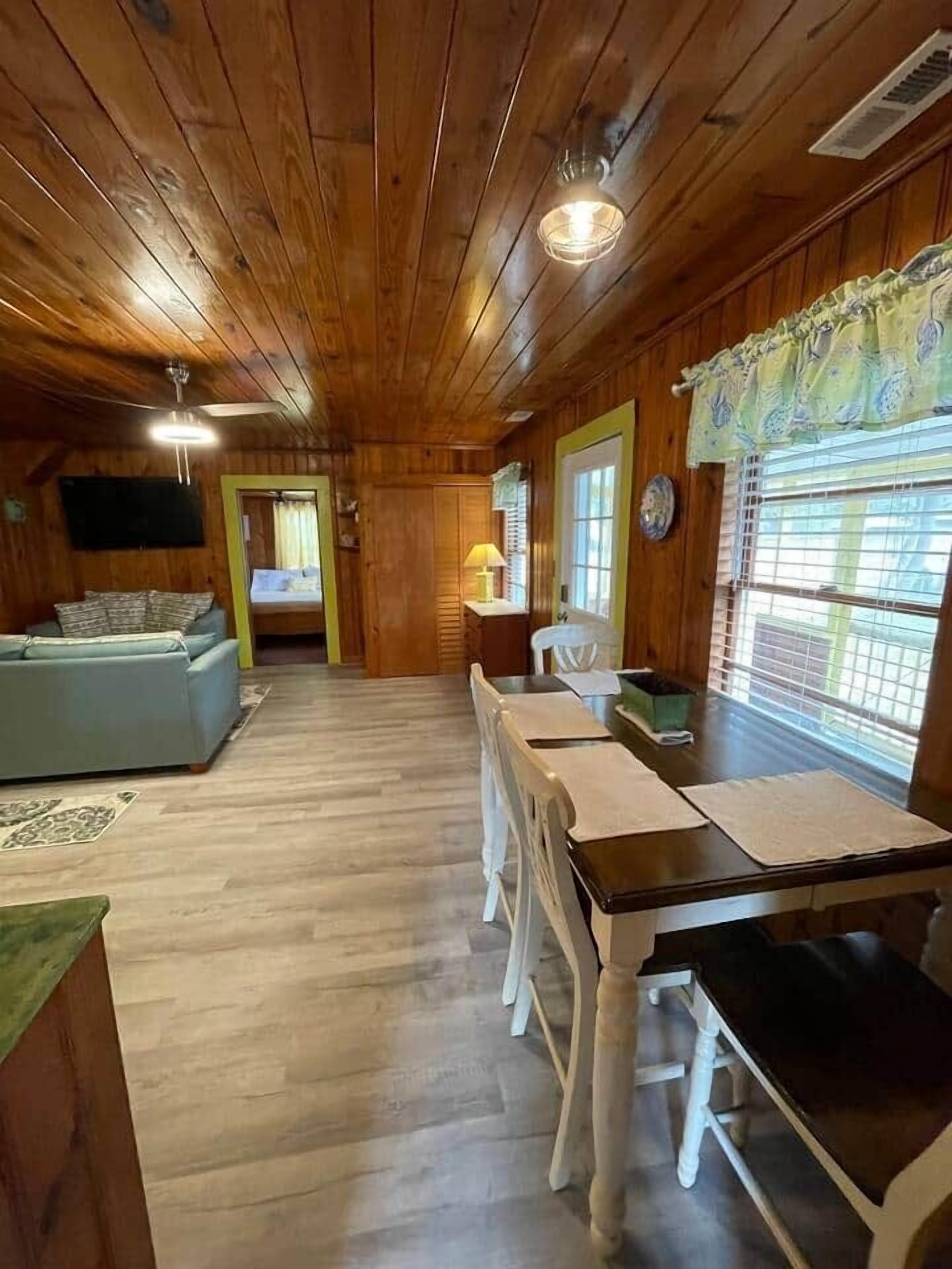 A wooden cabin interior with a dining table, chairs, a sofa, a lamp, and a window with blinds.