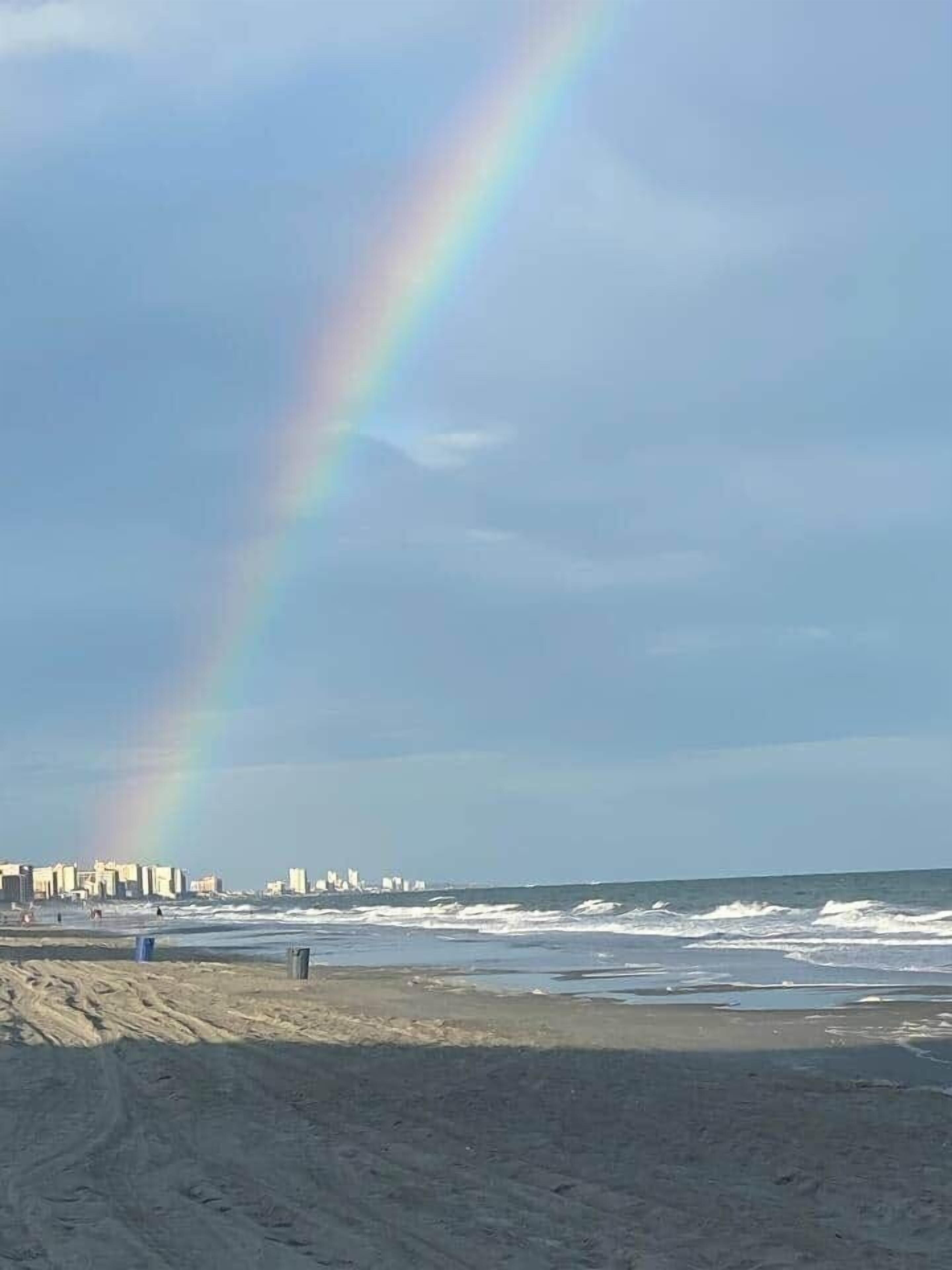 En strand med en regnbåge, en stadsbild i bakgrunden och vågor.