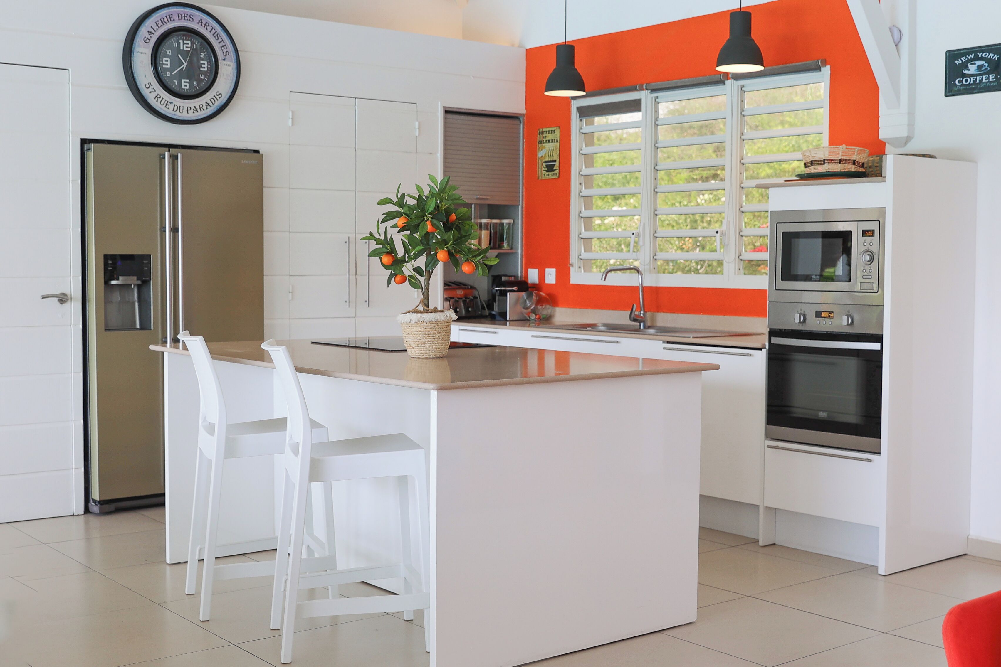 Une cuisine moderne avec des meubles blancs, un îlot central doté de chaises, un réfrigérateur, un four et une horloge murale.