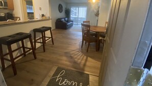 A modern kitchen with wooden countertops and bar stools, a dining area with a wooden table and chairs, and a living room with a sofa and a large window.