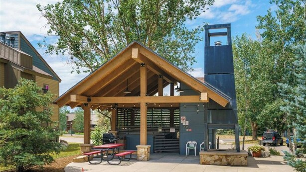 A covered outdoor seating area with picnic tables and a barbecue grill, surrounded by trees and a building with a chimney.
