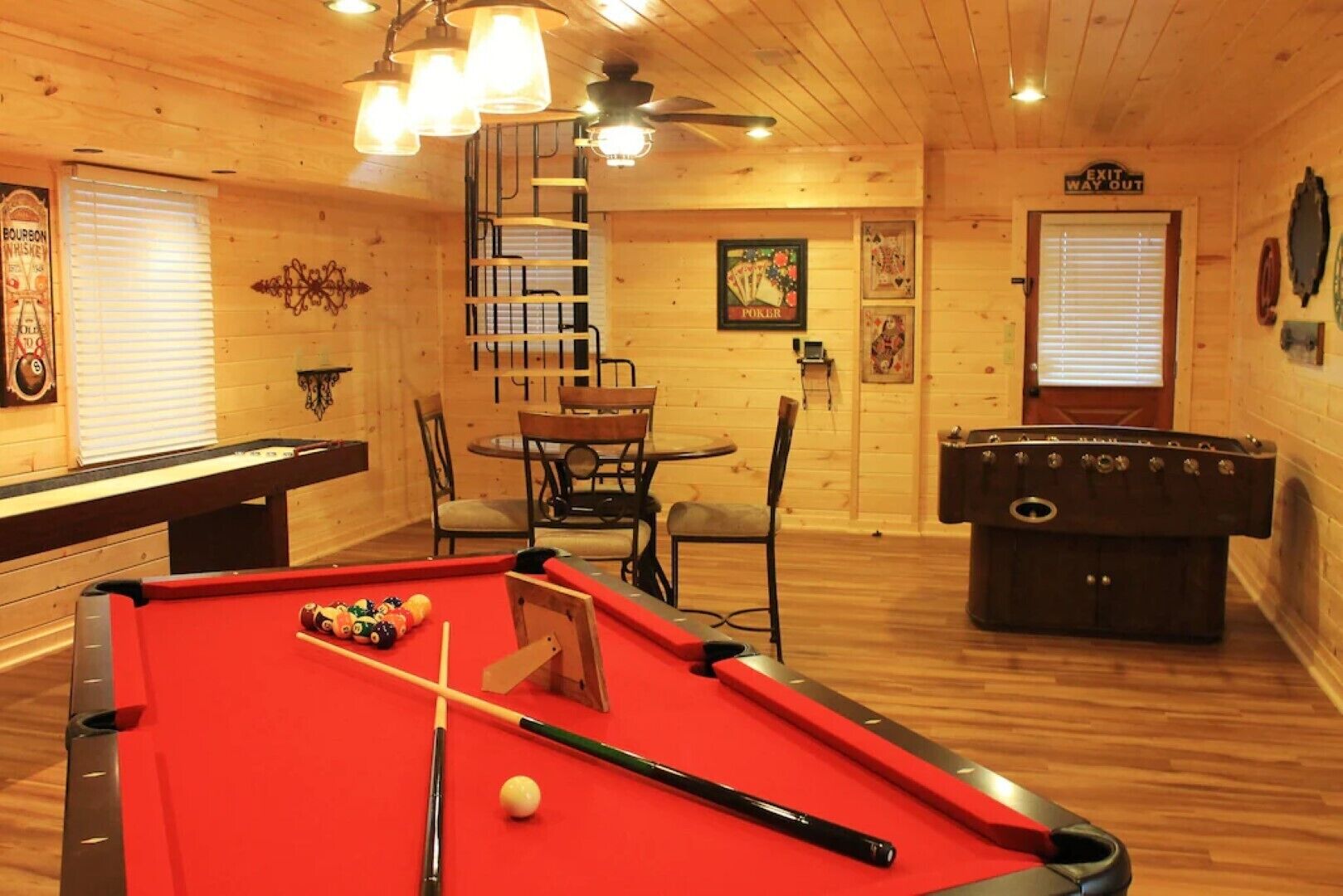 Een pooltafel met een rode viltbekleding, een biljartkeu en een witte poolbal. Rechts staat een tafelvoetbaltafel en boven een pokertafel hangt een bord. De ruimte heeft houten wanden en een plafondventilator.