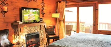 A cozy cabin room with a stone fireplace, a television, leather chairs, and a large bed.