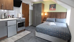 A bedroom with a bed, a nightstand, a wall-mounted TV, and a kitchen area with appliances. - Victoria House - Deluxe Studio by CovStays (Coventry)