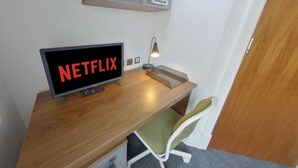 A small workspace with a wooden desk, a chair, a desk lamp, and a monitor displaying "NETFLIX". - Victoria House - Deluxe Studio by CovStays (Coventry)