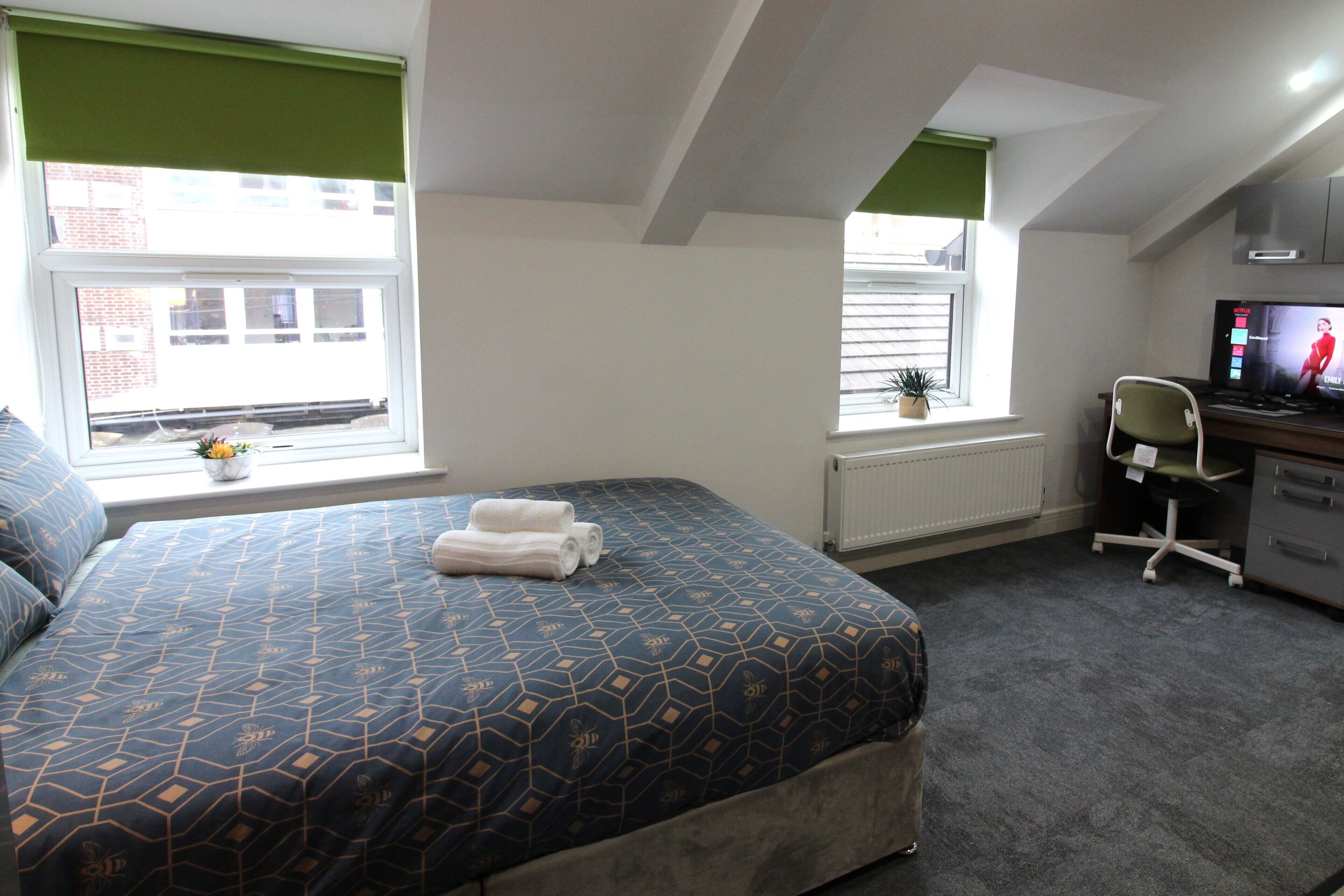 A bedroom with a bed, a desk, a chair, a radiator, and a window with blinds.