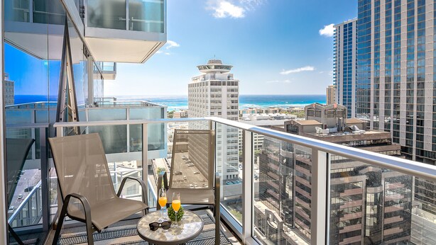 A balcony with a view of the ocean and cityscape, featuring a chair and table with drinks.