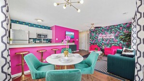 A modern kitchen with white cabinets, a pink accent wall, and a dining area with teal chairs and a white table. - The Pink Flamingo | Bold & Vibrant By The Beach (Myrtle Beach)