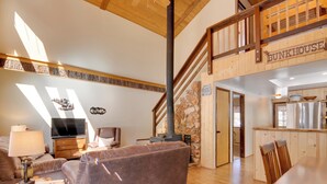 A spacious living room with a large brown sofa, a television, and a stone wall. A wooden staircase leads to a kitchen area with a sign reading "BUNKHOUSE." - Unique Forest Cabin with Deck: Ski, Hike, Fish! (Duck Creek Village)
