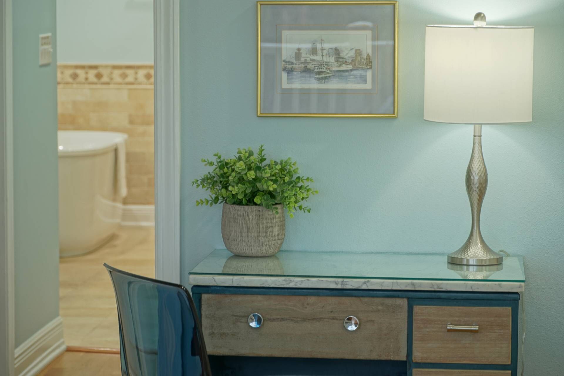 A bathroom vanity with a glass top, a potted plant, a framed picture, and a lamp.