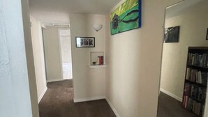 Um corredor com piso de carpete, uma parede branca com um quadro emoldurado, uma estante com livros e um espelho grande.