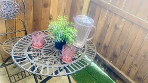 Une table ronde en métal, ornée d’une plante en pot et de deux carafes en verre, entourée de chaises en métal, le tout placé devant une clôture en bois.